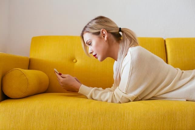 a young blonde woman is checking her phone while lying on the sofa